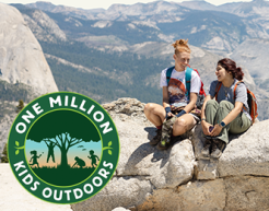 Two teens sit and chat on a large rock with a mountain range behind them. To the left is the logo for the One Million Kids Outdoors logo.