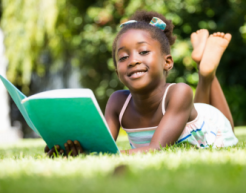 A smiling child reads a large book while lying in the grass