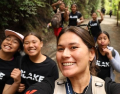 Ginny Ellis-Kirifi takes a selfie while hiking along a forest trail with six students, all wearing black shirts featuring the logo for the BLAKE nonprofit.