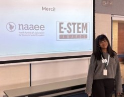 A young student poses for a photo in front of a projection screen, which displays the logos for NAAEE, the E-STEM Awards, and the word "Merci!"
