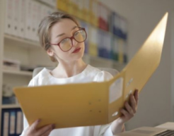A bespectacled person looks through a folder in a room with files filling the shelves behind them. 