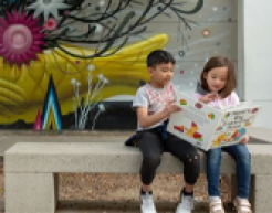 Two children sit next to one another on a bench, reading a large storybook, in front of an abstract mural