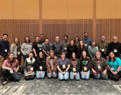 A group of people kneeling with a group standing behind, all wearing nametags around their necks, them on a carpeted floor with a wood paneled wall behind them