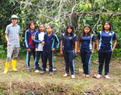 Wearing outdoor gear, six students pose with a teacher in front of dense trees.