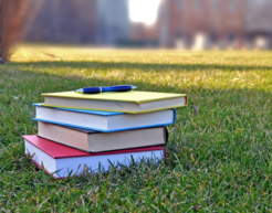 Four books stacked on a grass lawn with a pen on top of them; the background is a blurred university campus