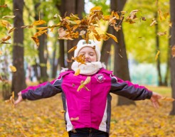 A young child in a jacket, scarf, and knitted hat throws a pile of autumn leaves up into the air.