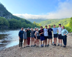 Twelve students from the James River Association's Upper James expedition pose for a group photo, standing along the riverbank of the James River. 