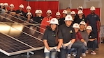Group photo of twenty people wearing hard hats next to a solar panel at a solar panel installation