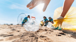 A hand reaches down to pick up a crumpled plastic bottle in the sand. 