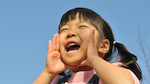 A smiling child with pigtails holds their hands up to shout.