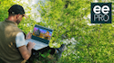 Person wearing hat an vest sits in the woods, looking at the NAAEE website on a laptop