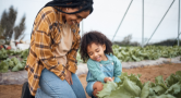 An adult kneels next to a garden with a young children touching the plant growing from the ground. 